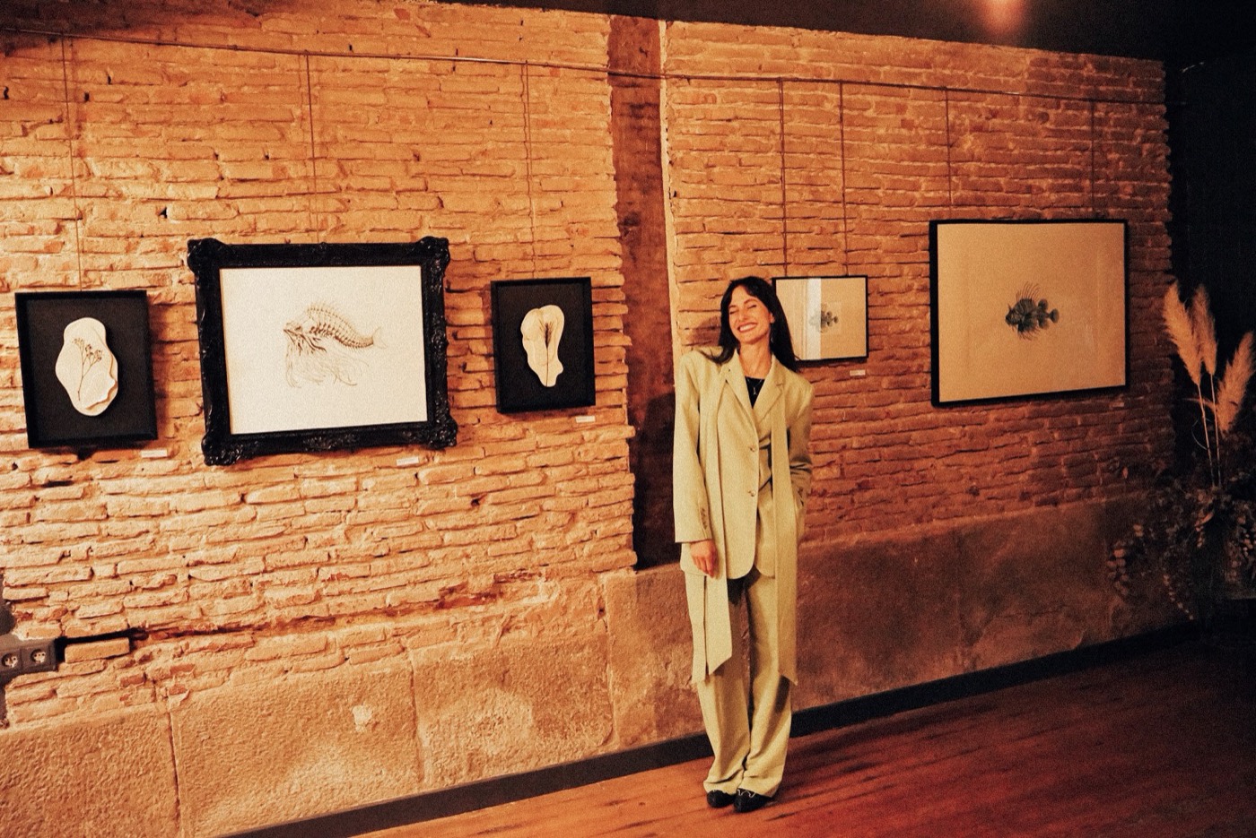 Valeria de pie junto a sus obras enmarcadas en una pared de ladrillo